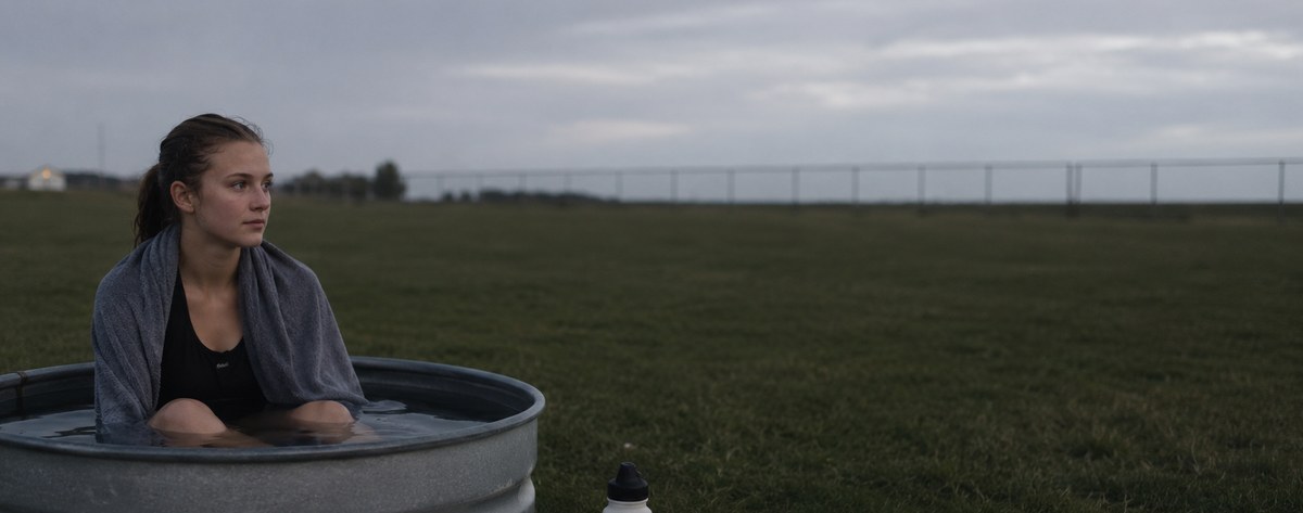 A high-school-age distance runner sitting in a plain galvanized stock tank of cold water on a practice field at dusk, towel over her shoulders, calm and unremarkable.