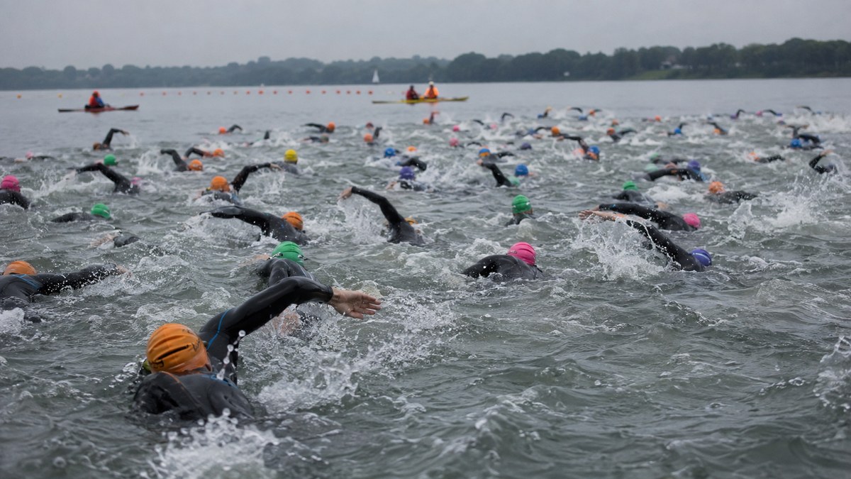A large pack of triathletes in black wetsuits and colored swim caps swimming freestyle at a mass open-water triathlon start, kayak safety escorts and a buoy line in the distance.