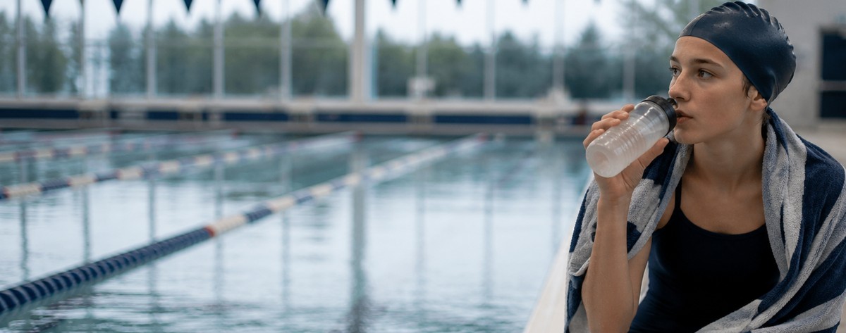 A high-school swimmer on the pool deck at dawn wrapped in a towel, eating a banana before early-morning practice.