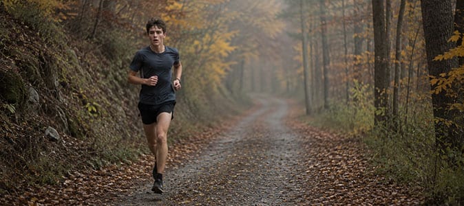 A high school cross-country runner alone on a wooded autumn trail, mid-stride, looking fatigued.